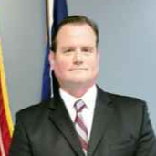 Supervisor Scott Musacchio brown suit standing in front of flag